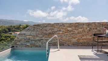 Una piscina de piedra con pared y escalera metálica, una mesa y sillas en una terraza con vistas al paisaje.