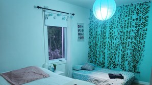 Ett sovrum med två sängar, ett fönster, en hängande lampa och en vägg med hängande växter.