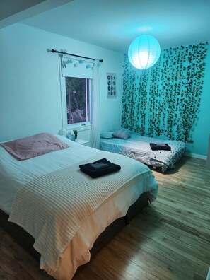 A bedroom with two beds, a window, a hanging light fixture, and a wall with hanging plants.