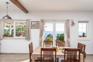 A dining area with a table and chairs, large windows with curtains, a pendant light, and a framed picture on the wall. - Villa Ole | Mediterranean Stone Villa with Pool, Jacuzzi (Jesenice)