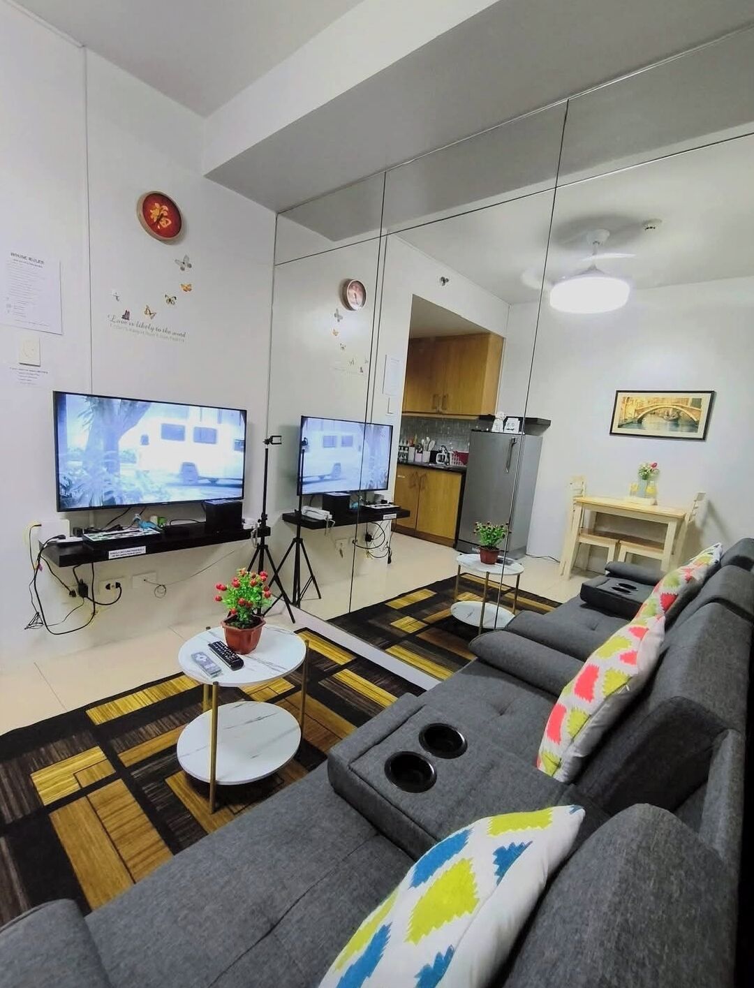 A modern living room with a sofa, two televisions, a small table with a plant, and a kitchen area in the background.