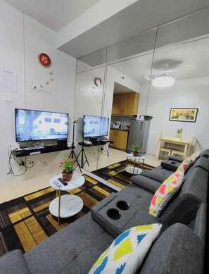 Ein modernes Wohnzimmer mit einer Couch, zwei Fernsehern, einem kleinen Tisch mit einer Pflanze und einer Küchenzeile im Hintergrund.