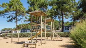 A wooden playground structure with a slide and climbing net, surrounded by sand and trees. - Village - Climatisé - 4p (3) (Le Bois-Plage-en-Ré)