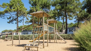 Una struttura giochi in legno con scivolo e rete da arrampicata, circondata da sabbia e alberi.
