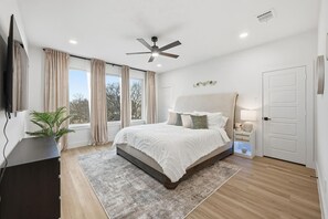 Ein Schlafzimmer mit einem Bett, einem Deckenventilator, einem Nachttisch mit Lampe, einer Pflanze und einem großen Fenster mit Vorhängen.