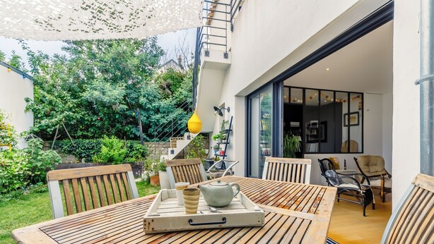 Un terrazzo in legno con tavolo e sedie, una lampada pendente gialla e vista su un giardino e un edificio.