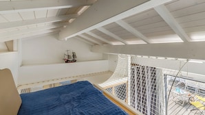 Una stanza con soffitto alto e tetto spiovente, un letto con copriletto blu, un modellino di nave su uno scaffale e una finestra zanzariera che si affaccia su un balcone.