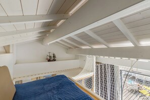 Ein Zimmer mit hohen Decken und geneigtem Dach, ein Bett mit blauer Bettdecke, ein kleines Schiffmodell auf einem Regal und ein mit einem Netz versehenes Fenster mit Blick auf einen Balkon.