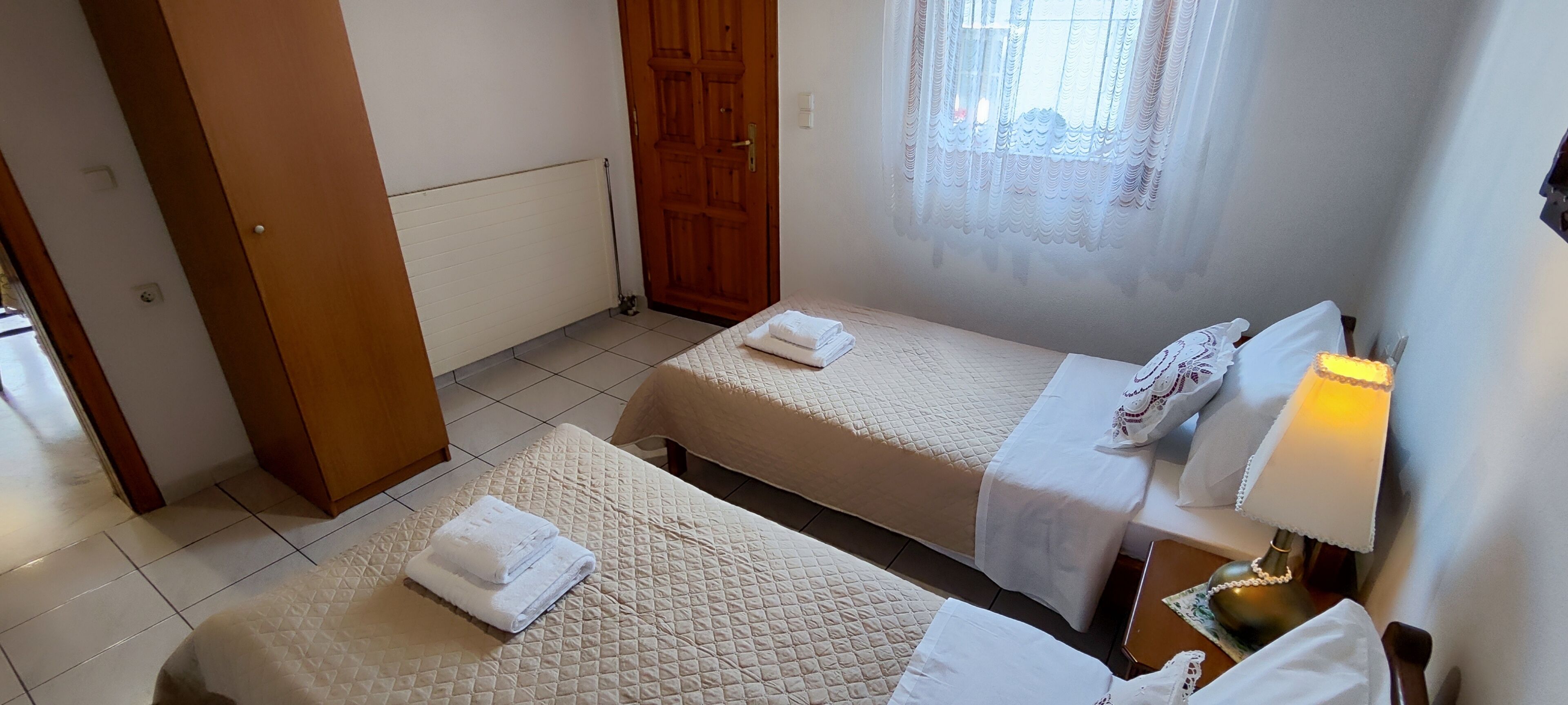 A hotel room with two beds, a wooden door, a radiator, a window with curtains, and a bedside lamp.