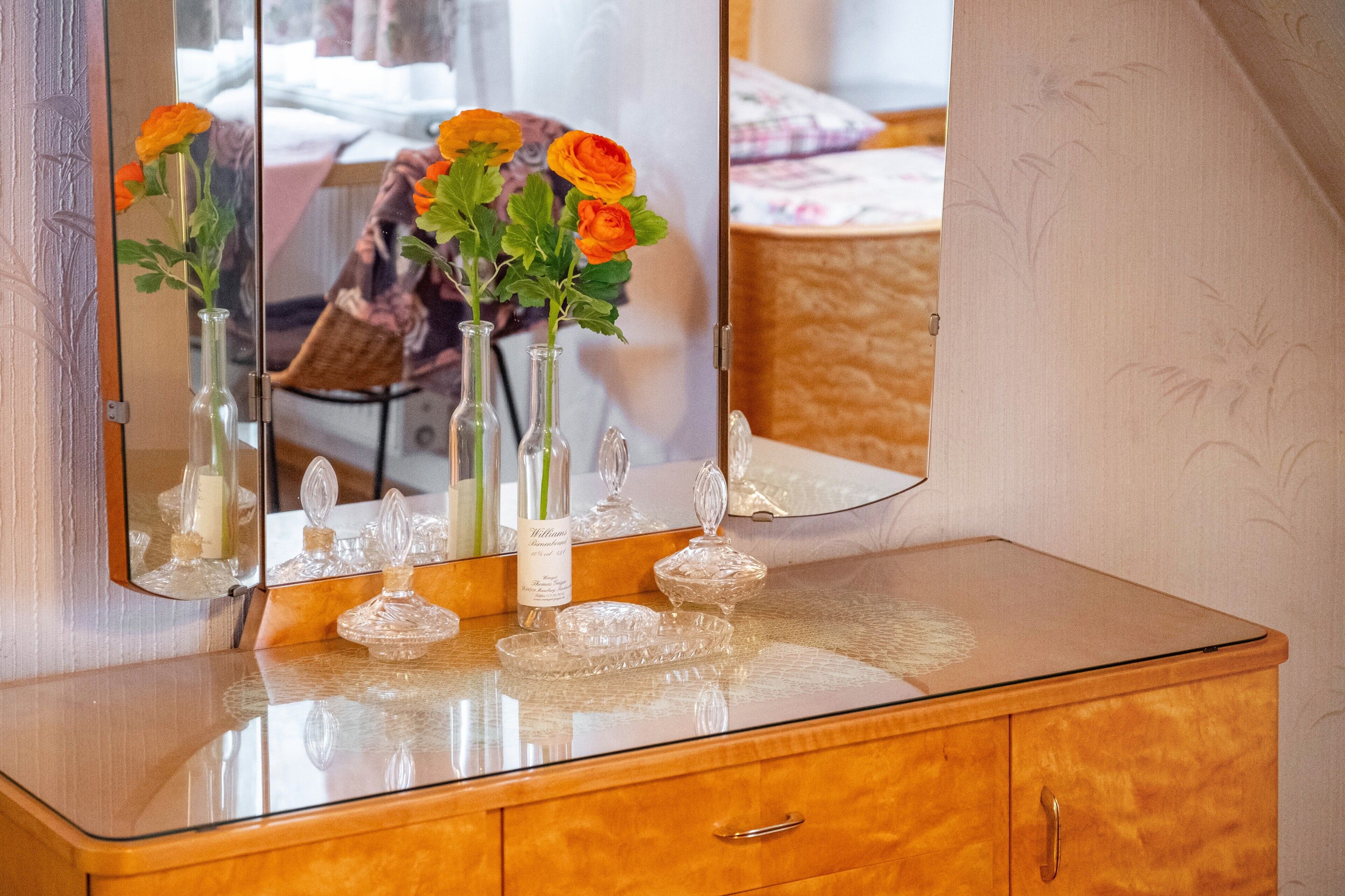Ett sminkbord i trä med en glasskiva som bordsyta, en spegel och tre genomskinliga glasvaser med orange blommor.