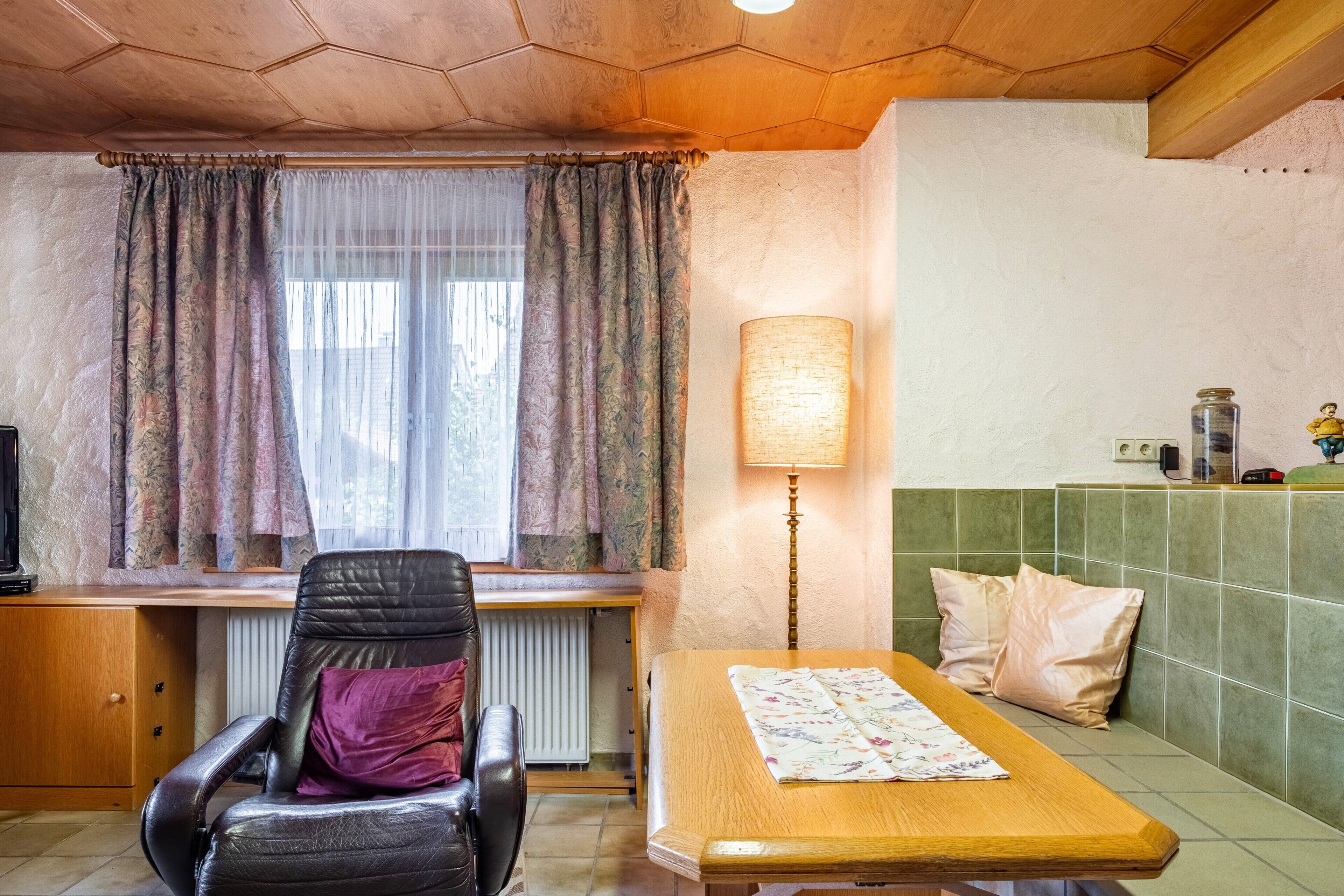 A room with a wooden desk, a leather chair, a table with a placemat, a lamp, and a green tiled wall.