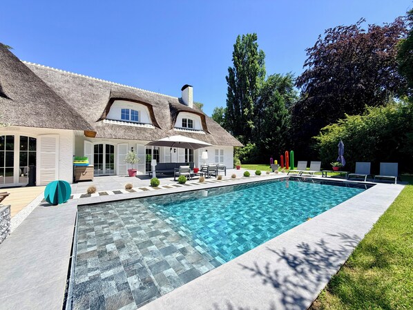 Une maison avec une piscine, un patio aménagé avec du mobilier d’extérieur et un toit de chaume.
