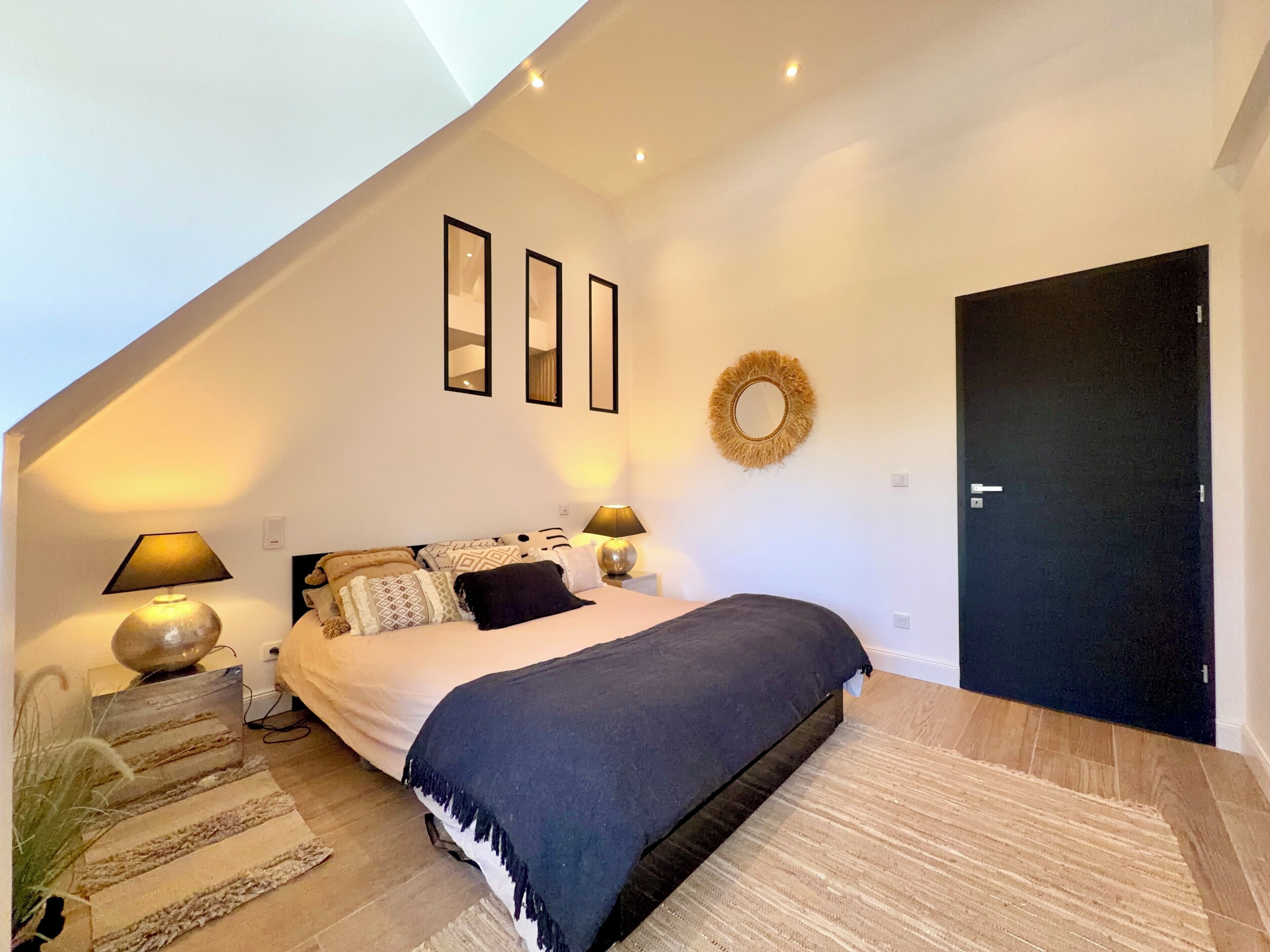 A bedroom with a bed, two bedside lamps, a mirror, a door, and a wall-mounted wreath.