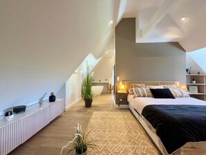 Un dormitorio moderno con techo inclinado, una cama grande, una mesita de noche y una planta en maceta, además de una bañera integrada.