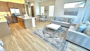 Eine moderne Küche mit einer zentralen Kochinsel, ein Essbereich mit Tisch und Stühlen, ein graues Sofa und ein gemustertes Teppich.