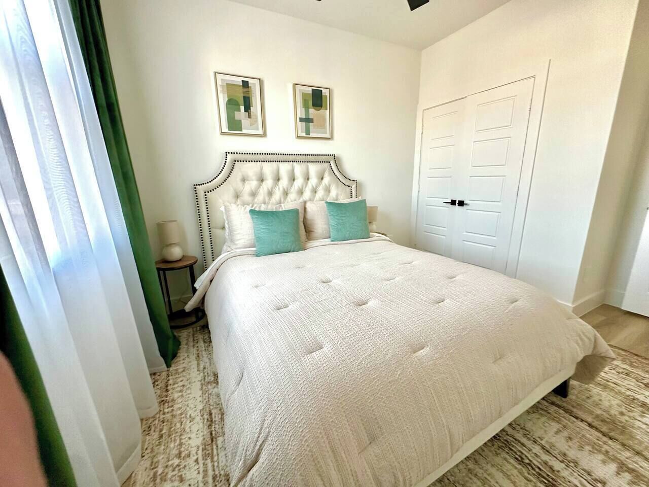 Une chambre à coucher avec une tête de lit capitonnée, un lit recouvert d’une courtepointe blanche texturée, deux coussins turquoise, une petite table de chevet avec une lampe et une porte dotée d’une poignée.