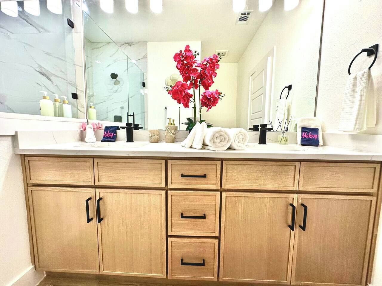 Un baño con tocador de mármol, cabina de ducha con paredes de vidrio, espejo y un jarrón con flores rojas.