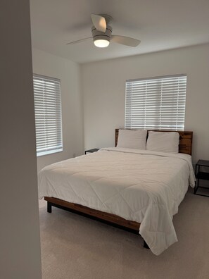 Ein Schlafzimmer mit einem Bett, zwei Fenstern mit Jalousien, einem Deckenventilator und einem Nachttisch.