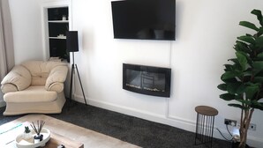 Un salon avec un canapé beige, un téléviseur à écran plat, une cheminée, une petite table ronde et une plante en pot.