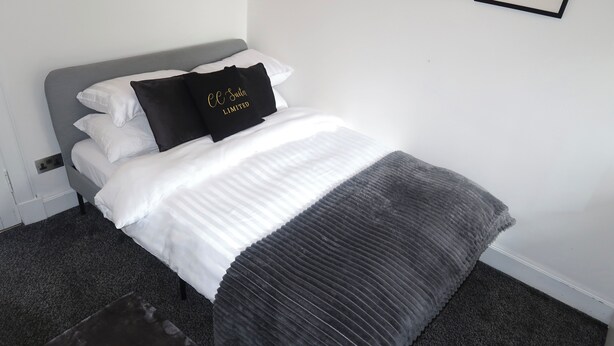 Un lit bien fait, avec une literie blanche et grise, un coussin noir orné d’un motif textuel et un tapis gris foncé.