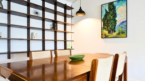 Un comedor con una mesa de madera, sillas, un tazón verde, un estante adosado con objetos decorativos, y un cuadro.