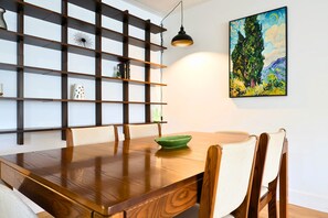 Un coin repas avec une table en bois, des chaises, un saladier vert, une étagère murale ornée d’objets décoratifs et un tableau.