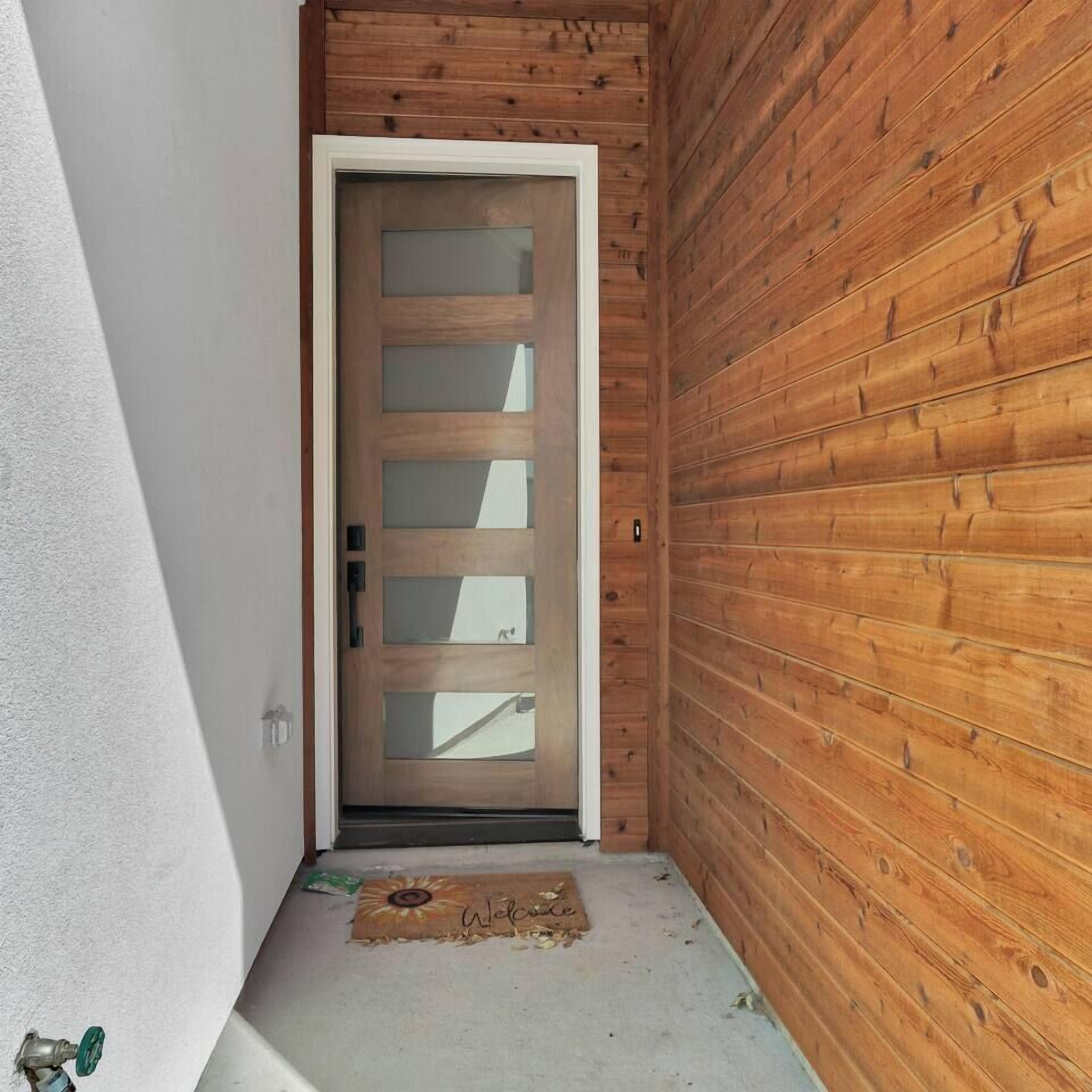 Uma porta de madeira com painéis de vidro jateado, um tapete de entrada com um desenho de sol e uma parede branca ao lado da porta.