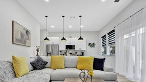 Uma sala de estar moderna com um sofá modular cinza, almofadas amarelas e pretas, uma mesa de centro de madeira com um vaso, luminárias pendentes e uma cozinha ao fundo.