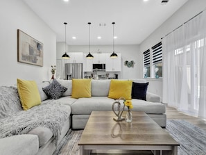 A modern living room with a grey sectional sofa, yellow and black cushions, a wooden coffee table with a vase, pendant lights, and a kitchen in the background. - Bright Dallas 3BR | Rooftop, Garage, Backyard - Holiday Home (Dallas)