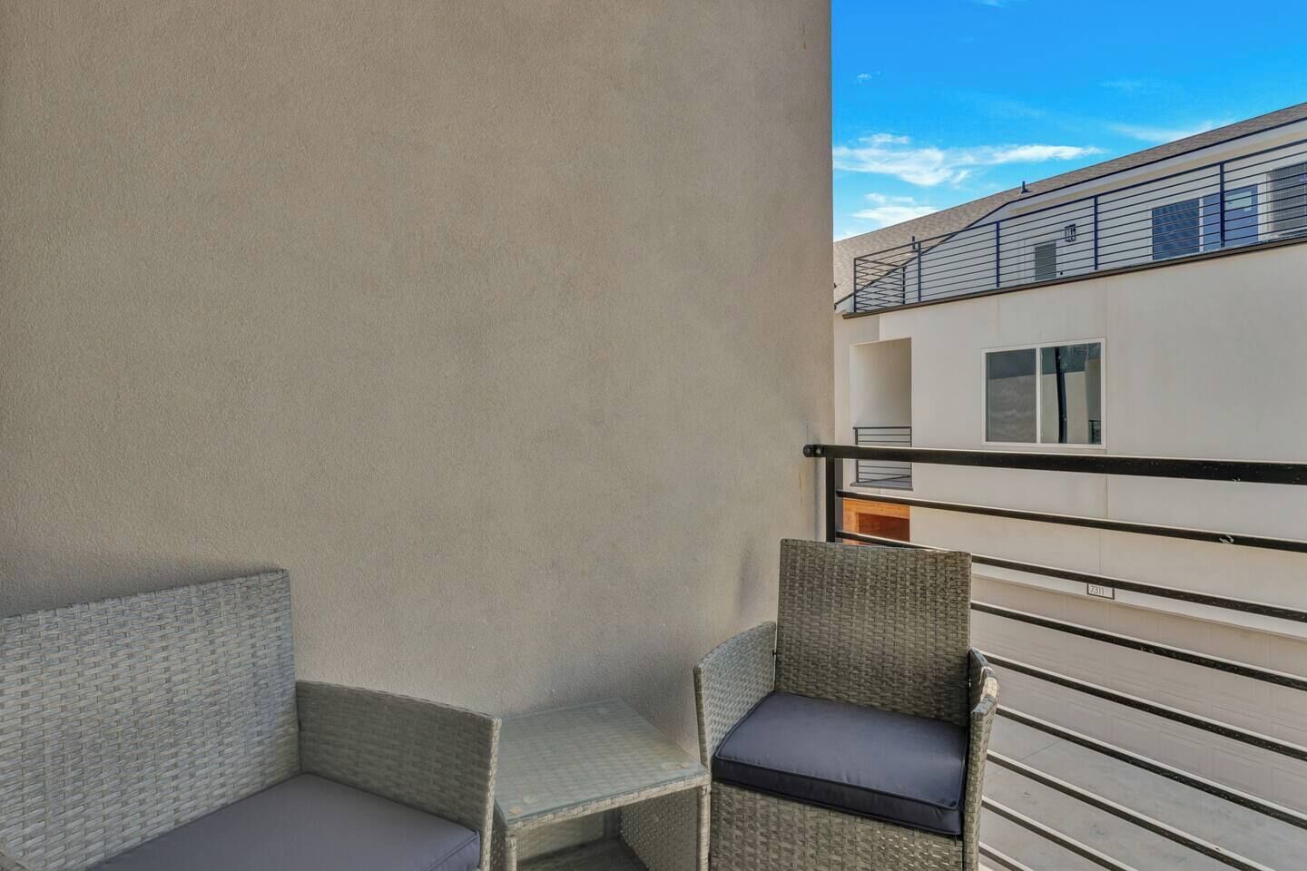 Een balkon met rieten meubels, een metalen hekwerk en uitzicht op omliggende gebouwen.