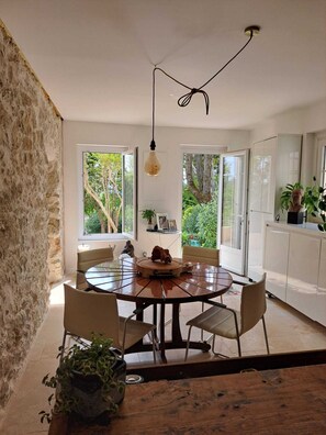 A dining area with a round table, chairs, a hanging light fixture, and large windows with a view of greenery.