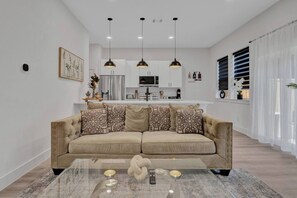 Ein modernes Wohnzimmer mit einem beige-farbenen Sofa, einem Glastisch und einer Küche im Hintergrund.