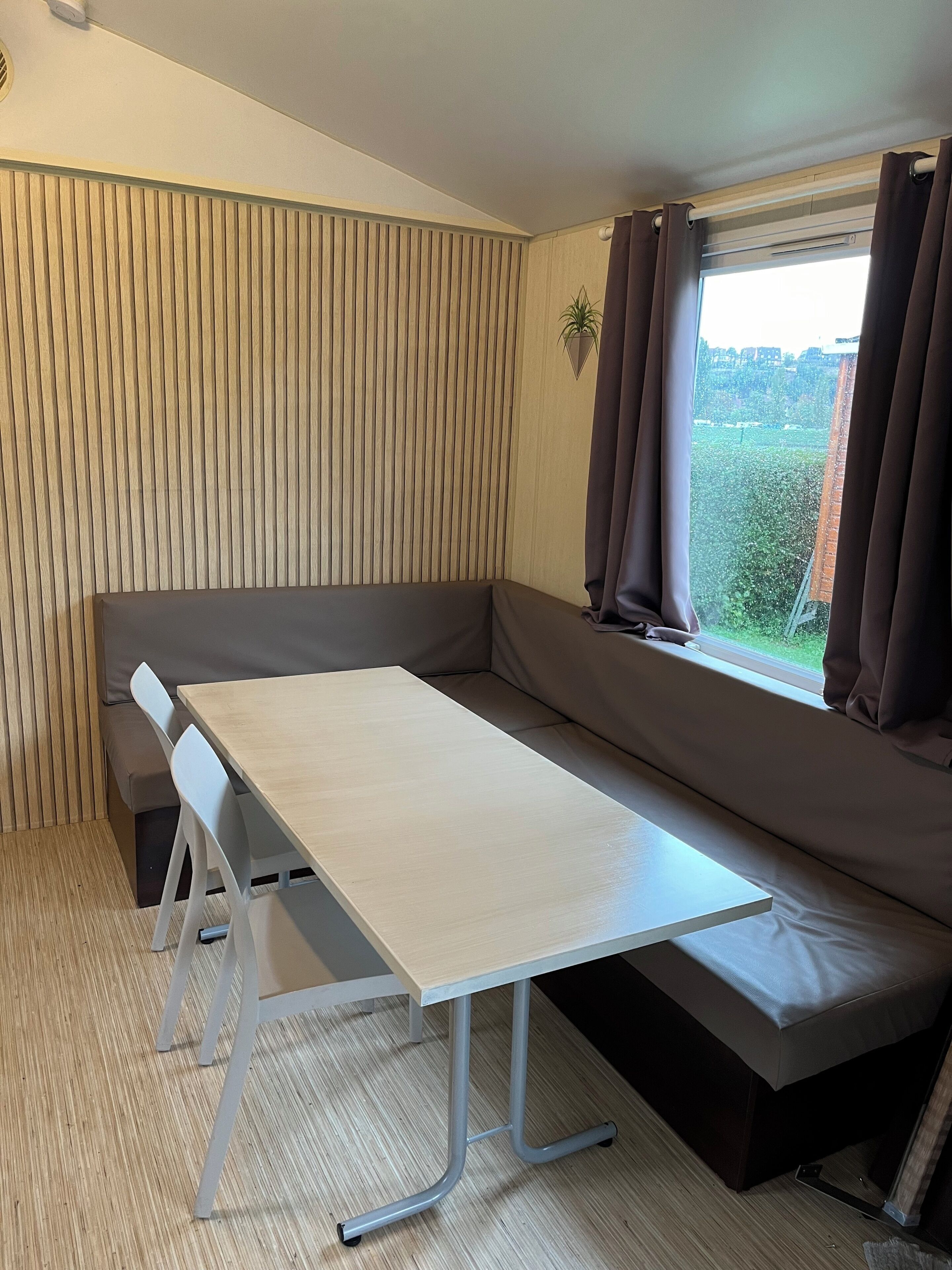 Une pièce avec un mur lambrissé en bois, un banc, une table avec des chaises et une fenêtre avec des rideaux.