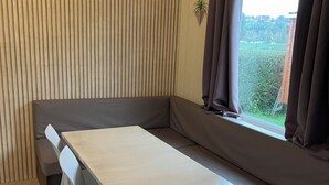 Una habitación con pared de listones de madera, un banco, una mesa con sillas y una ventana con cortinas.