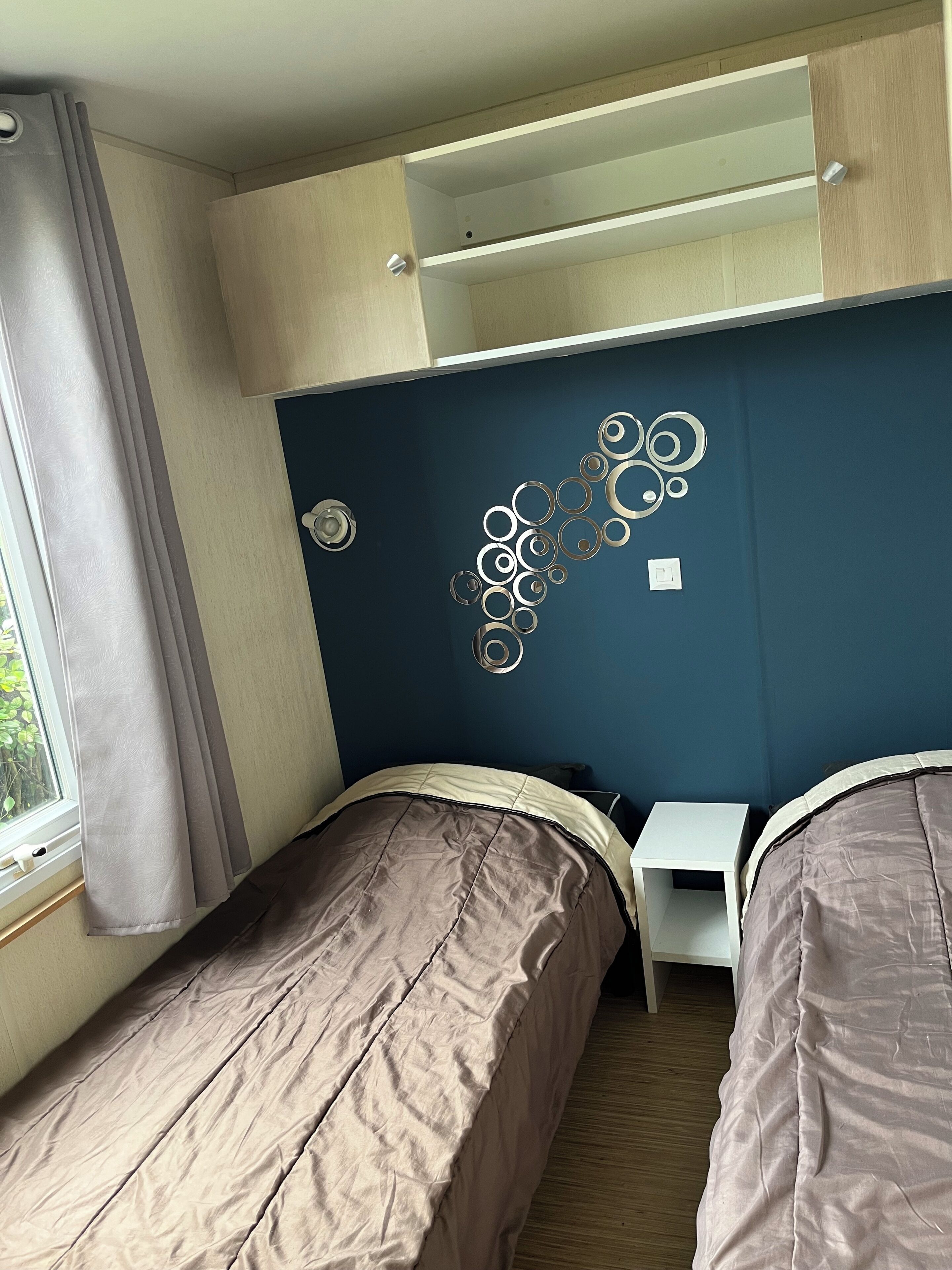 Une chambre avec deux lits, une fenêtre avec des rideaux, une étagère fixée au mur et un élément décoratif en métal.