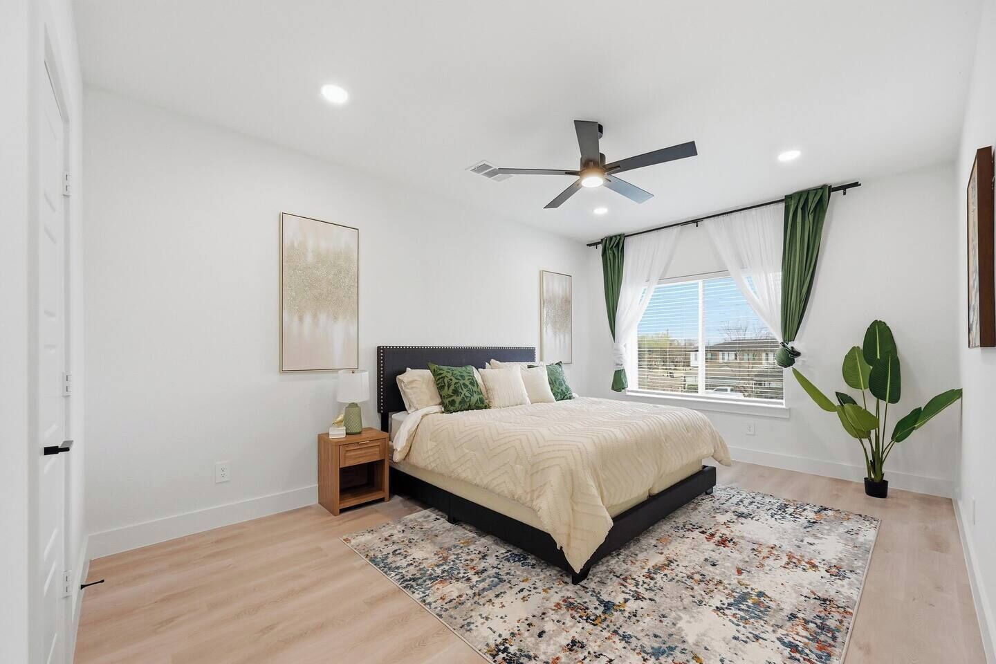 Un dormitorio con cama, ventilador de techo, mesita de noche, ventana con cortinas y una planta en maceta.