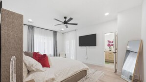 Een moderne slaapkamer met een bed, een plafondventilator, een televisie, een spiegel en een badkamer die door een open deur zichtbaar is.