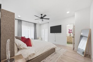 Ein modernes Schlafzimmer mit einem Bett, Deckenventilator, Fernseher, Spiegel und einem durchgehenden Türrahmen einsehbaren Badezimmer.