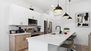 Een moderne keuken met witte kasten, roestvrijstalen apparatuur, een groot eiland met barkrukken, hanglampen en een minimalistisch kunstwerk aan de muur.