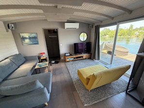 Ein modernes Wohnzimmer mit einer Couch, einem Fernseher und Blick auf eine Holzterrasse und Wasser.
