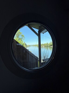 Ein rundes Fenster mit Blick auf ruhiges Wasser, einen Holzzaun und Bäume.