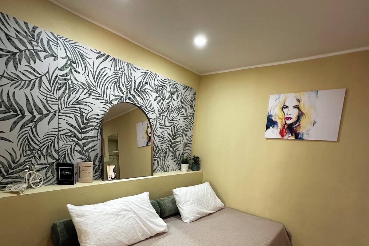 Une chambre à coucher avec un lit, des oreillers, un miroir, un mur orné d’un motif de feuilles et un tableau.