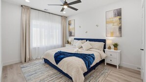 Ein Schlafzimmer mit einem Bett, einem Nachttisch, einem Deckenventilator, einem Fenster mit Vorhängen und Kunstwerken an der Wand.