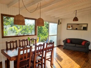 Un comedor con una mesa y sillas de madera, un ventanal con vistas a zonas verdes, un sofá gris y una lámpara colgante.