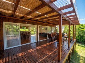 Una terraza cubierta de madera con un ventanal que da a una habitación, mobiliario exterior y vista a follaje.