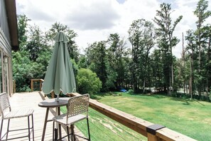 Un patio exterior con una mesa y sillas, una sombrilla verde y vistas a un jardín cubierto de césped y árboles.