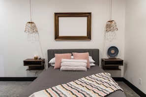 Ein Schlafzimmer mit Bett, Nachttischen, einem an der Wand befestigten Bilderrahmen und Pendelleuchten.