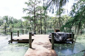 Eine Holzstege führt zu einer überdachten Sitzgelegenheit mit Blick auf ein sumpfiges Gebiet mit Bäumen und Wasser.
