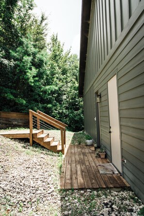 A wooden deck leading to a house with a door and a window, surrounded by a gravel area and greenery.