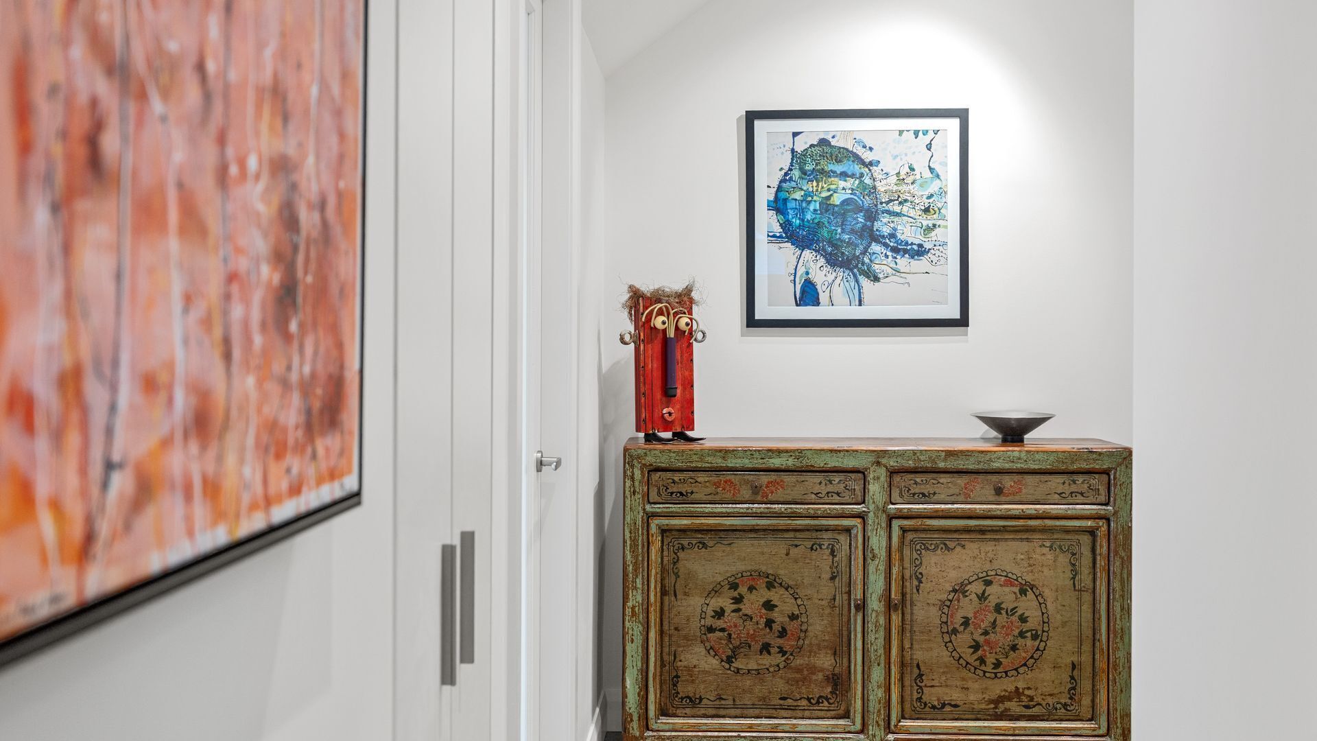 A hallway with a white door, a rustic wooden cabinet with floral designs, and a framed abstract painting on the wall.